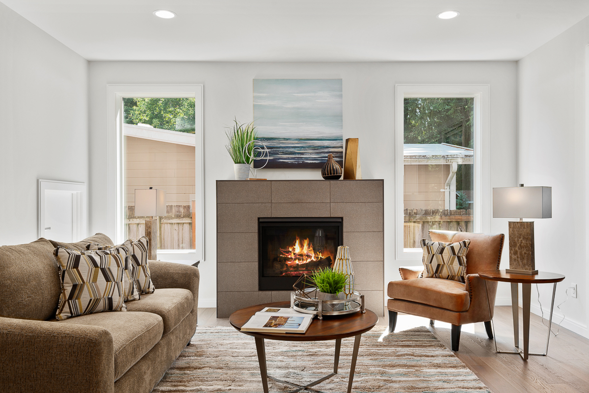 Sala de estar con chimenea, confortable sofá, sillón, mesas redondas, plantas y grandes ventanales con vistas al exterior.