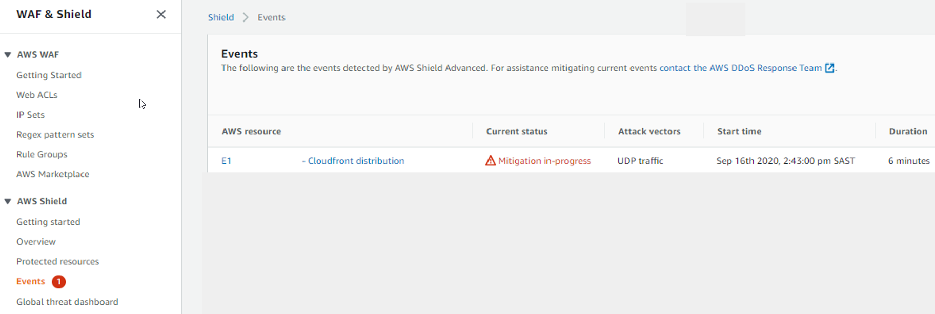 The AWS Shield console left navigation pane has the Events selection highlighted in red, with a numeral 1 beside it, inside a red circle. The Events page is open, and shows a single row in the events list. The row lists an AWS resource of type CloudFront distribution. The Current status field contains a triangular red icon next to the words Mitigation in progress. The Attack vectors status field contains UDP traffic. The Start time field contains Sep 16th 2020, 2:43:00 pm SAST. The Duration field contains 6 minutes.
