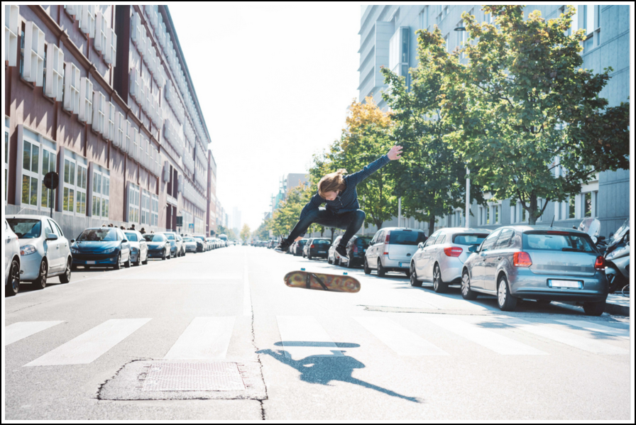 Persona che fa un'acrobazia su uno skateboard in mezzo a una strada cittadina tra macchine parcheggiate.